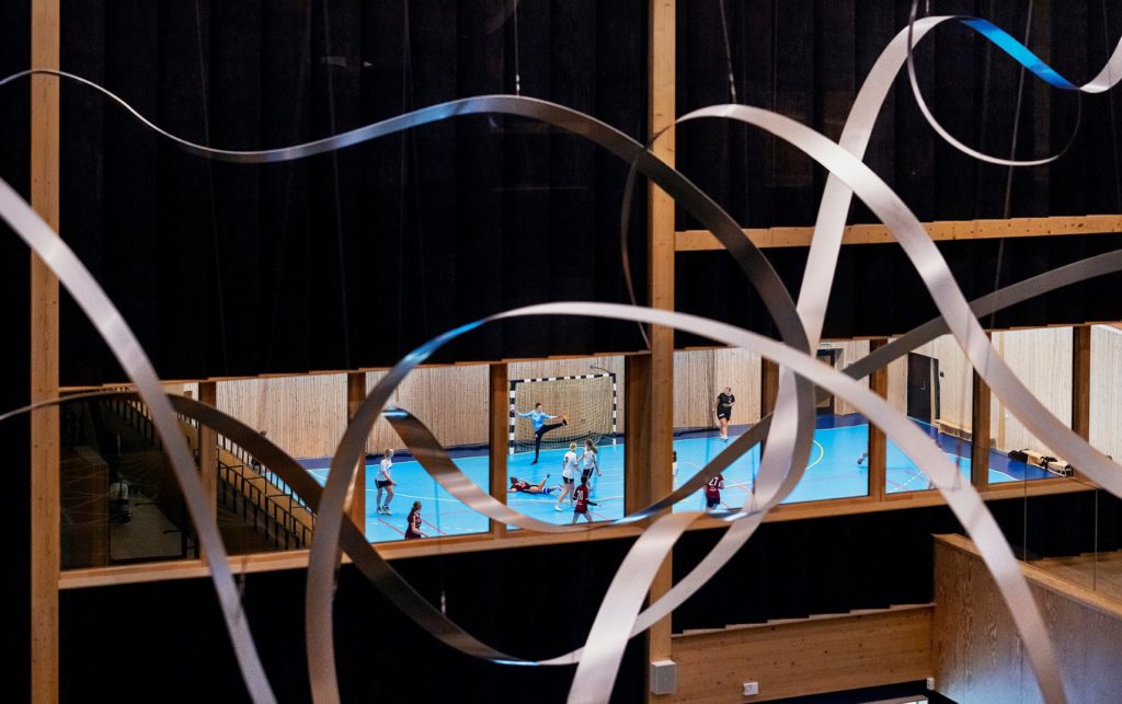 I förgrunden, konstverket Moves av formade stålband. I bakgrunden, sporthall med blått golv, handbollspelare.