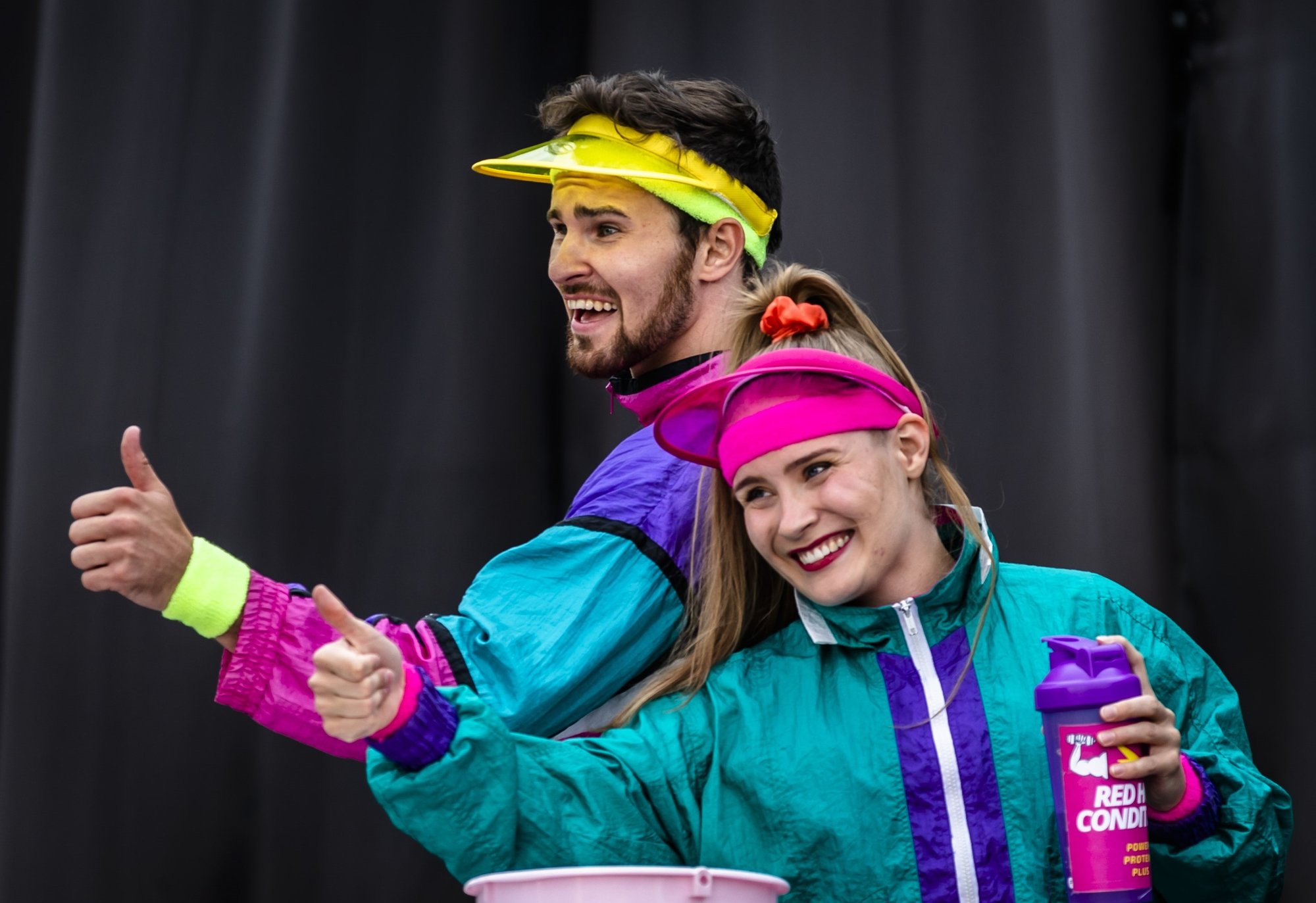 Två cirkusartister, en man och en kvinna, gör tummen upp i färgglada overaller