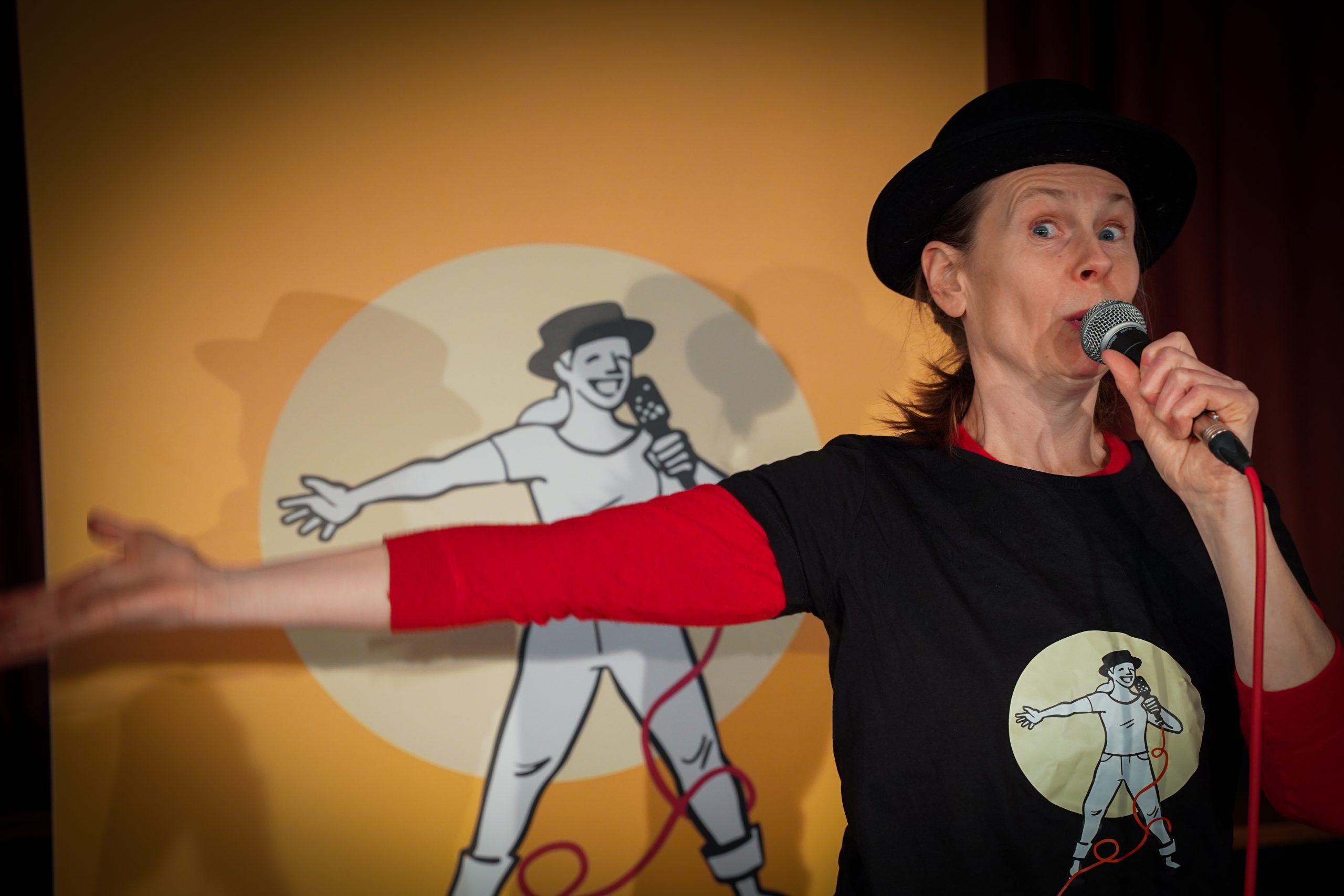 Michaela står framför en ritad bild av henne själv med höger arm rakt ut och håller i en mikrofon. Hon har en svart hatt på huvudet samt en röd tröja med en svart t-shirt med tryck på sig.