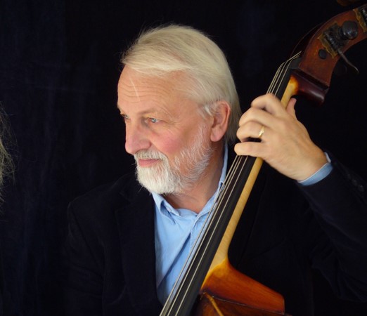 Man med vitt skägg och hår iklädd mörk kavaj och blå skjorta spelar på en ståbas