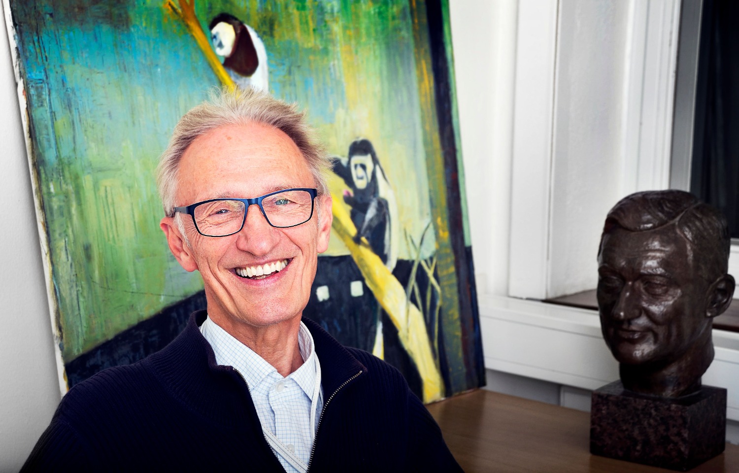 Professor Töre Theorell i vit skjorta, glasögon och en svart rock ler stort och står framför en färgglad målning med pandor som klättrar på en trädstam. Till höger syns en byst i svart material.