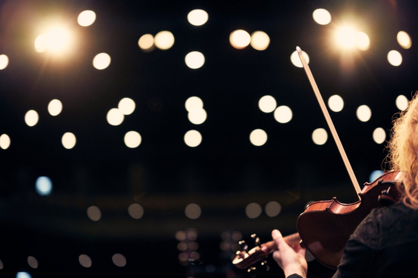 Lysande punkter a scenstrålkastare och en fiol och stråke i förgrunden som någon spelar på . En hand syns hålla fiolen.