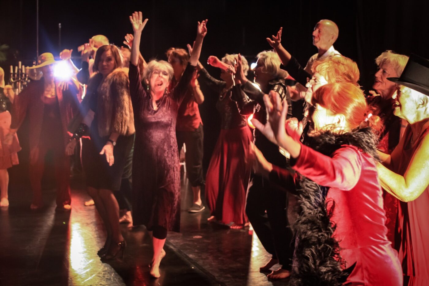 Äldre människor dansar i festliga klänningar på ett scengolv. En kvinna i lila klänning i mitten sträcker upp armarna i luften