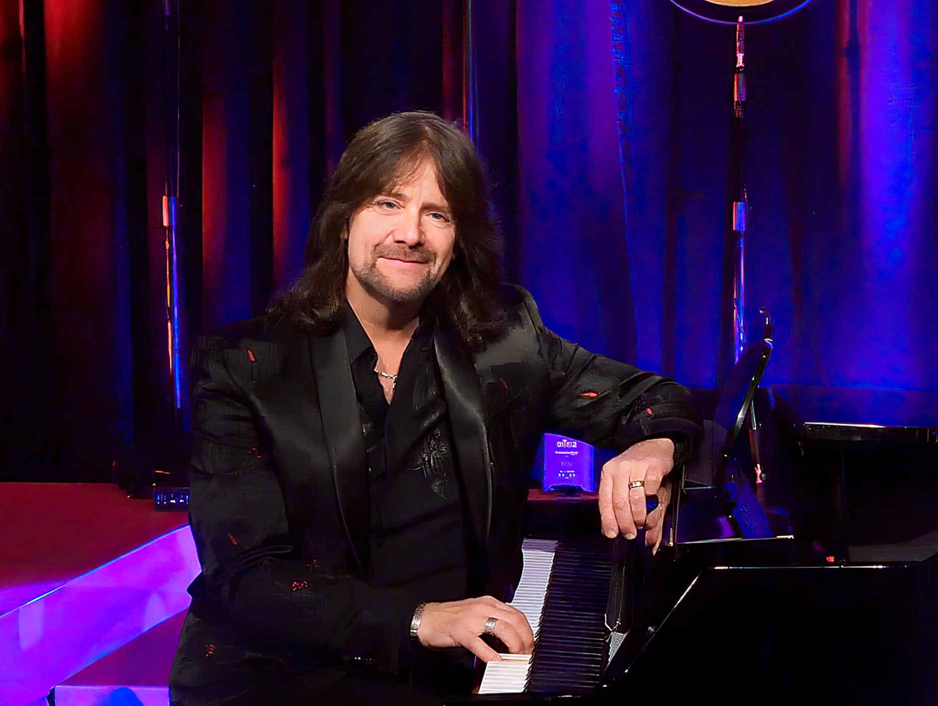 Robert Wells sitter vid flugeln med ena handen på pianotangenterna och ler mot oss. Han har långt hår och svart kostym.