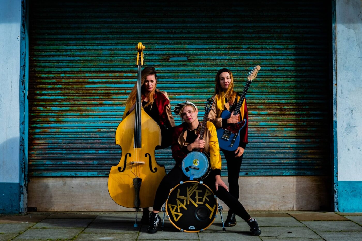 Tre kvinnliga musiker står bakom sina instrument som är ståbas, baskaggetrumma, banjo och gitarr. Scenkläderna går i blått och gult och lite rött med svarta tajta byxor. De står framför en vägg av rostig blå korrugerad plåt.