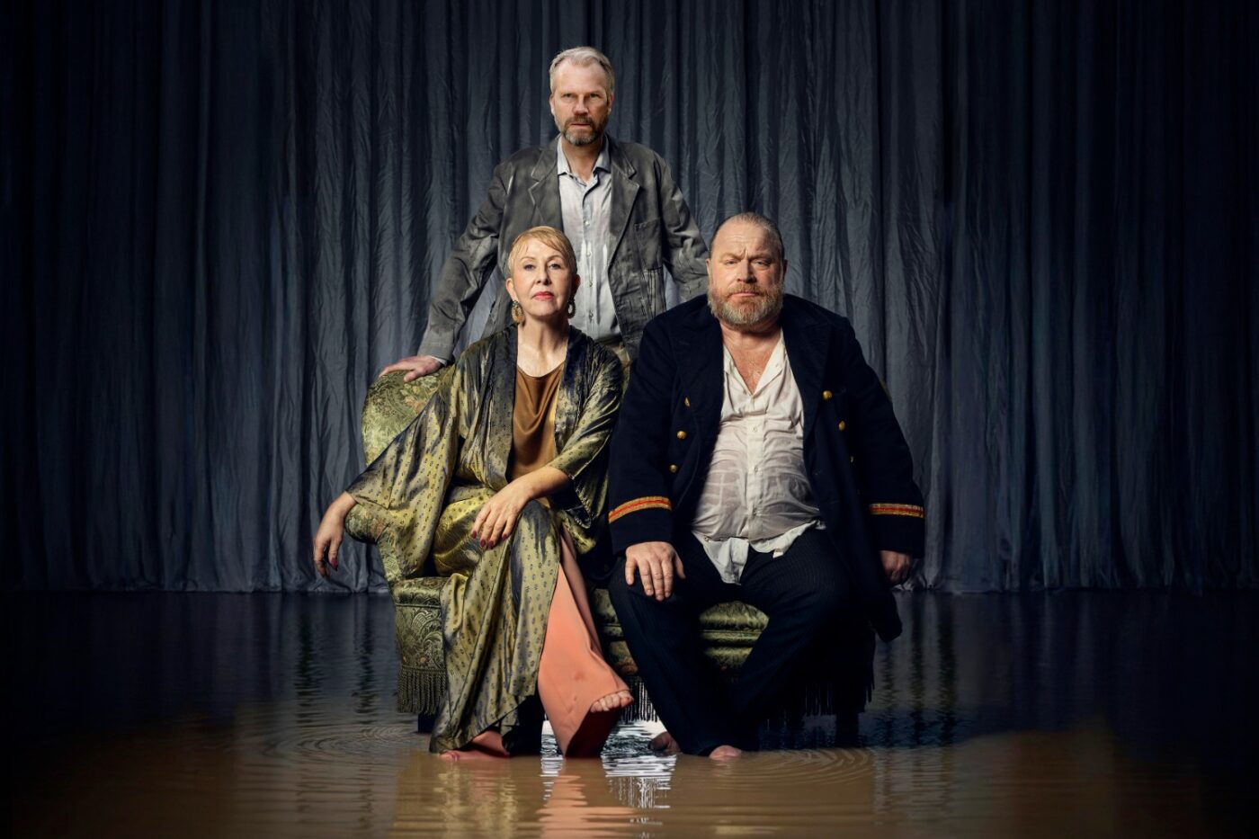 Tre skådespelare på en scen där golvet är täckt av brunt vatten. En stående man och en sittande man och en kvinna.