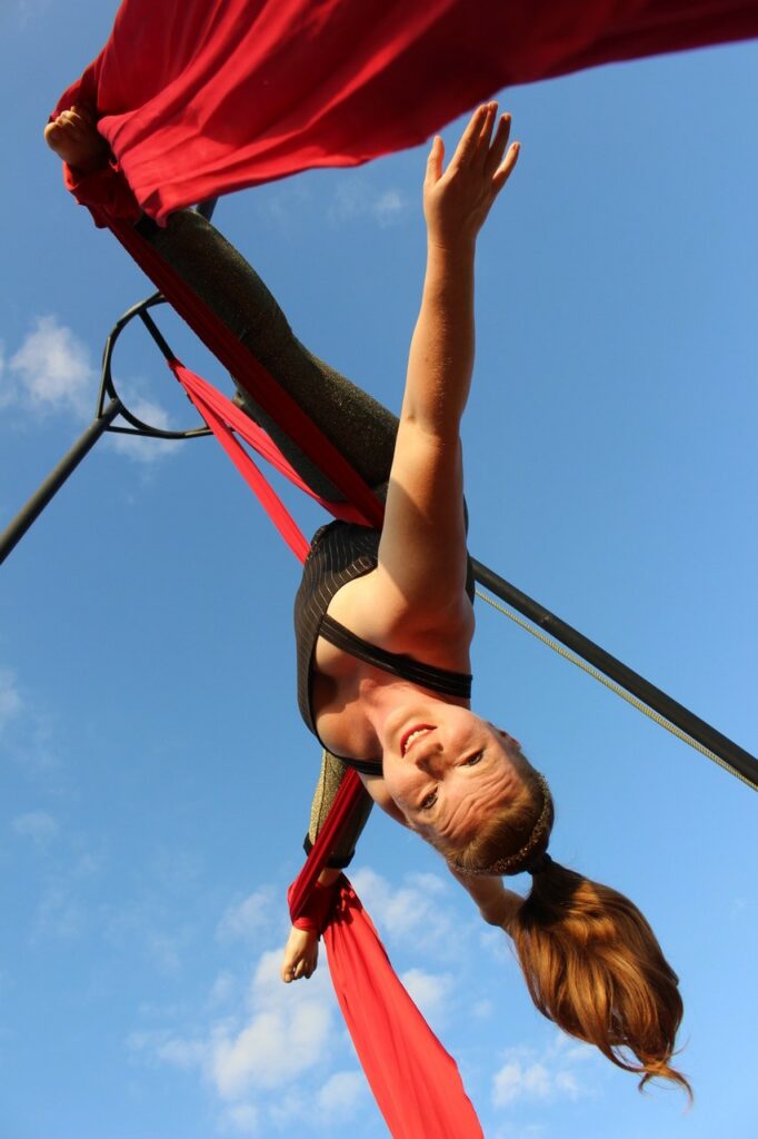 Luftakrobat hänger i upp och ned i ett rött tyg i en ställning utomhus mot en blå himmel och tittar ned mot oss.