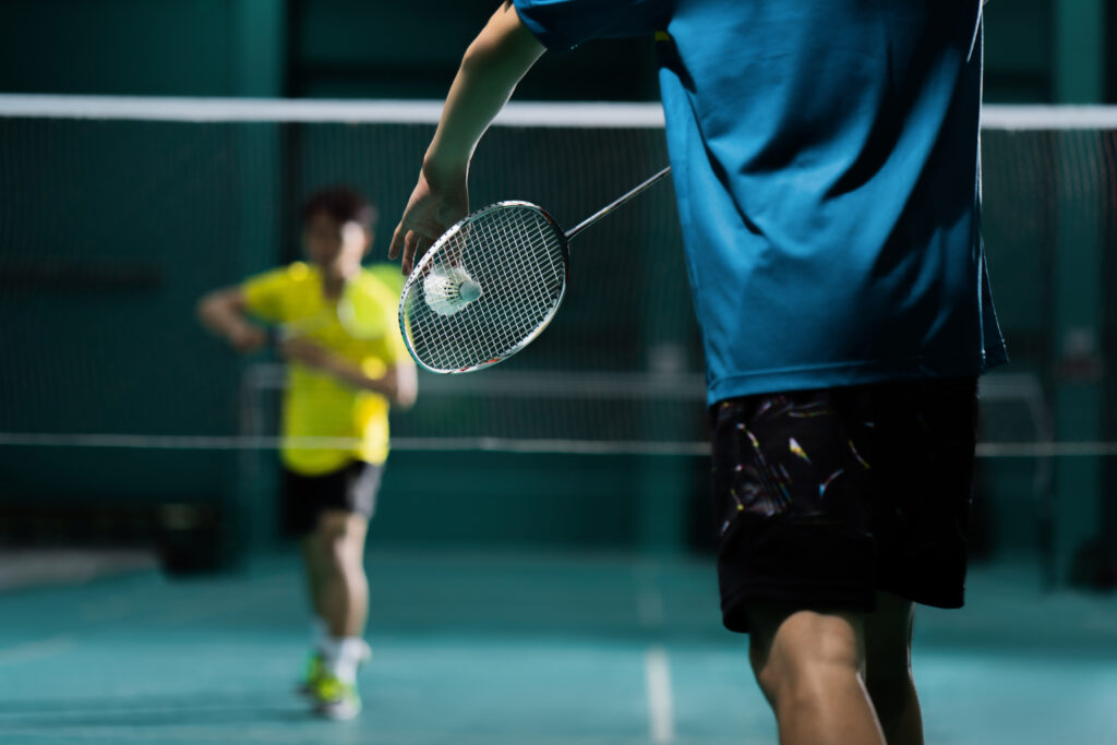 Två personer spelar badminton inomhus. Personen i förgrunden, klädd i blå tröja, slår precis till en fjäderboll med sitt racket. I bakgrunden syns motståndaren i gul tröja, oskarpt i fokus. En nät skiljer spelarna åt.
