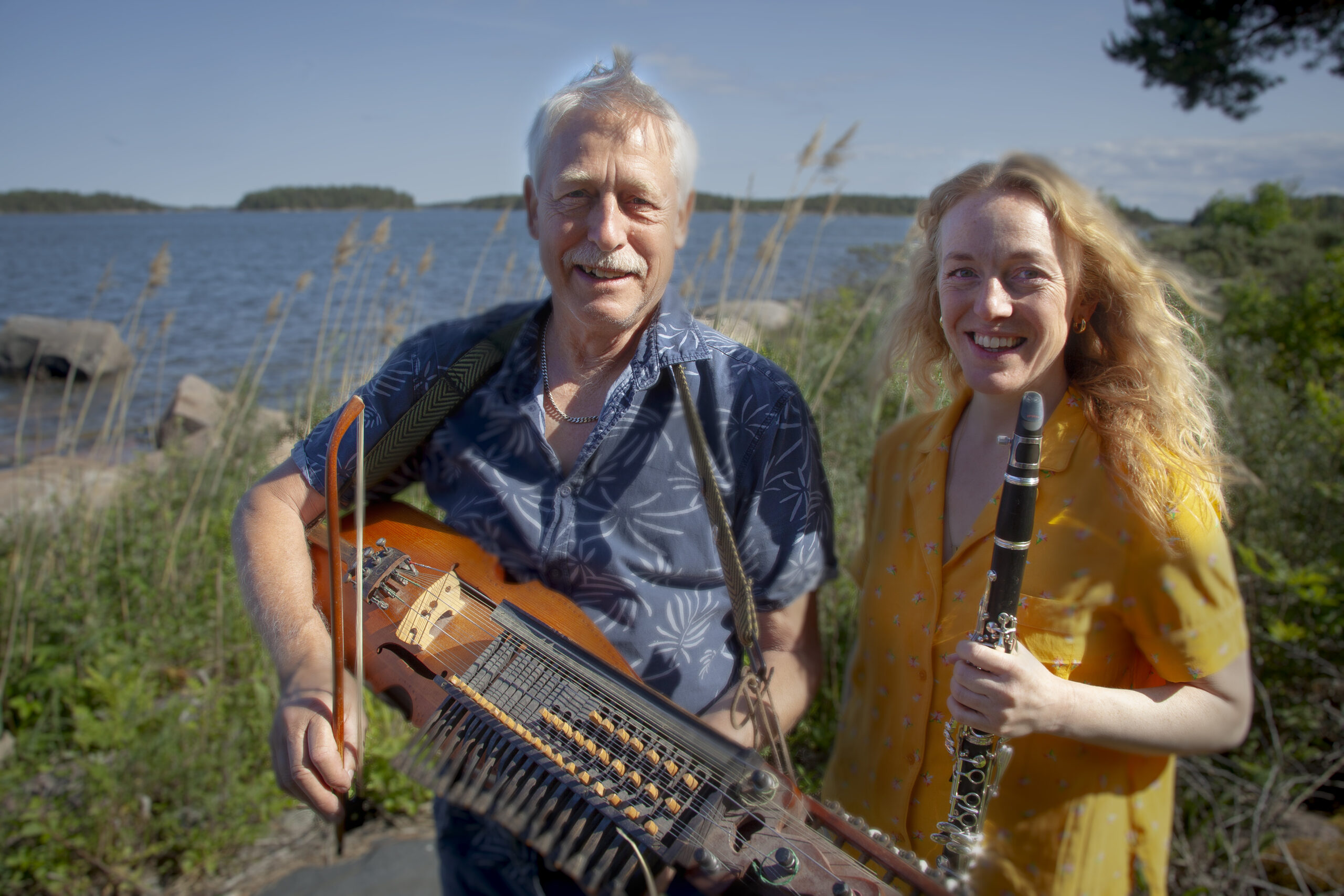 Man i blå skjorta med nyckelharpa och kvinna i gul klänning med klarinett står i solen framför landskap med sjö och gräs