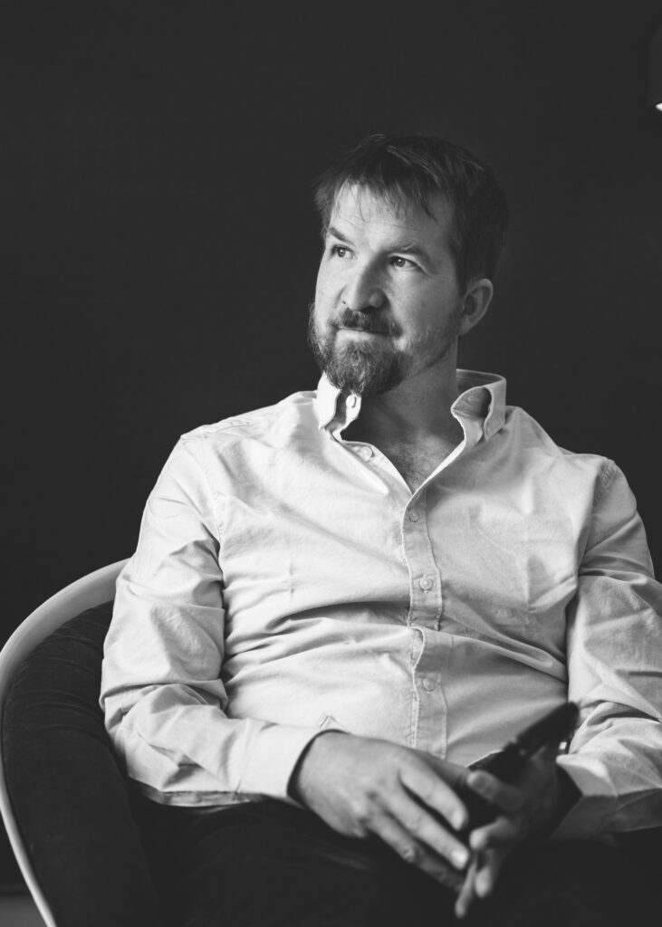 Svartvit porträttbild av Koreografen Robin Jonsson sittande på en stol och har en vit skjorta